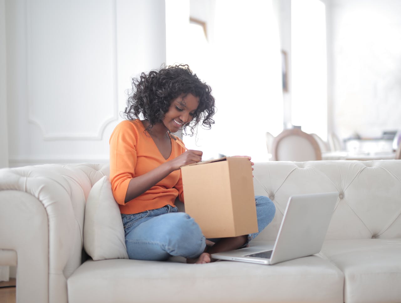 Side view of curly haired cheerful black woman opening carton box while sitting on sofa with laptop