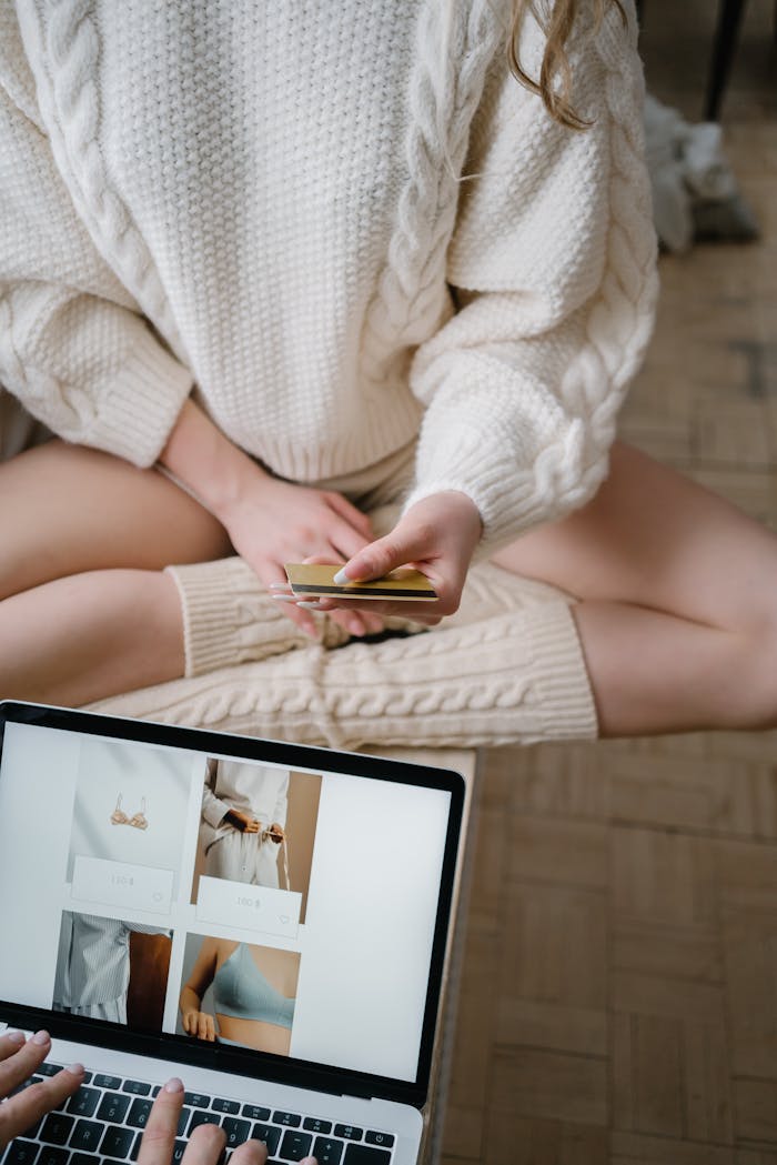 A woman in cozy attire shopping online using a laptop and credit card indoors.